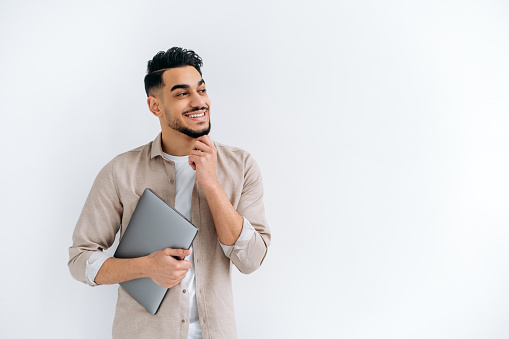 Positive handsome arabian or indian guy in stylish casual clothes, freelancer or student, holding a laptop, standing on a white isolated background, looking away, smiling happily. Copy space concept