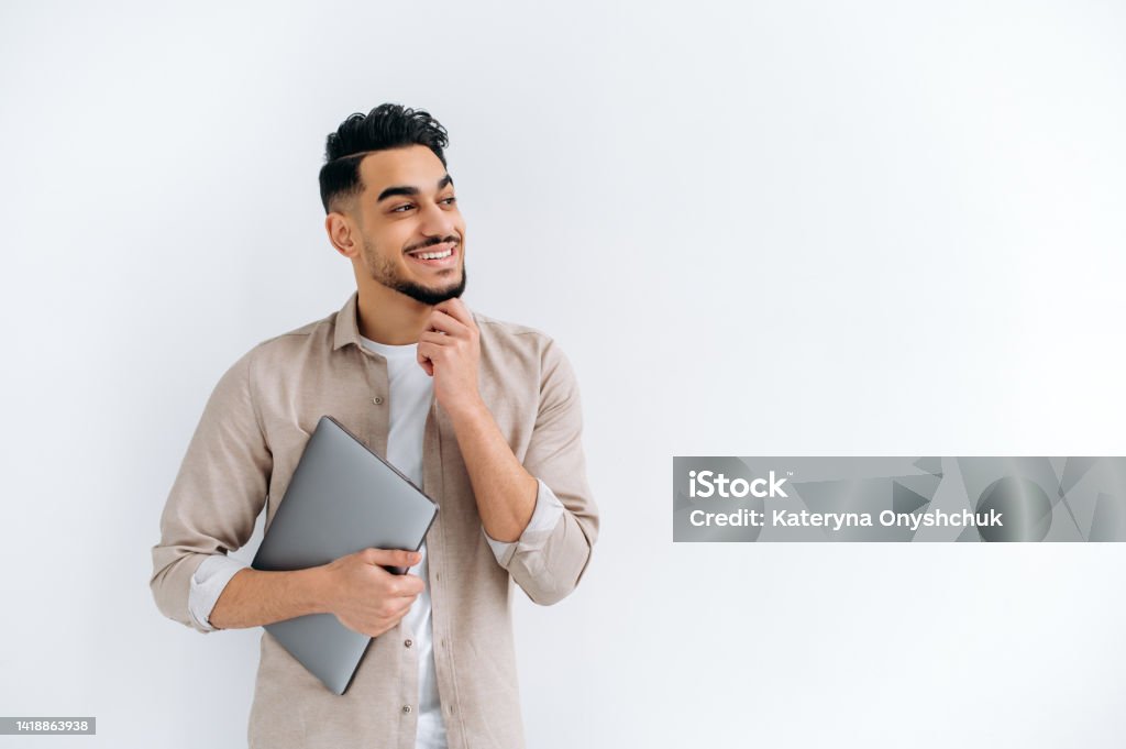 Positive handsome arabian or indian guy in stylish casual clothes, freelancer or student, holding a laptop, standing on a white isolated background, looking away, smiling happily. Copy space concept Student Stock Photo