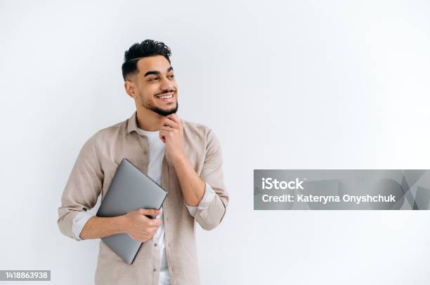Positive Handsome Arabian Or Indian Guy In Stylish Casual Clothes Freelancer Or Student Holding A Laptop Standing On A White Isolated Background Looking Away Smiling Happily Copy Space Concept Stock Photo - Download Image Now