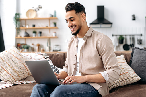 Modern successful smart indian or arabian freelancer, IT specialist, sits on a comfortable sofa in the living room, uses a laptop, works online, browsing the internet, social networks, smiles happily