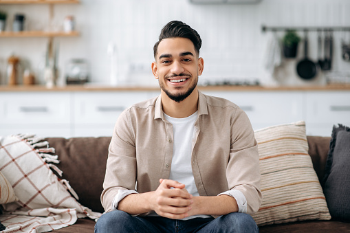 Portrait of an attractive handsome indian or arabian guy in a stylish casual wear, sitting on sofa at home in a living room, in a stylish interior, looking at the camera, smiling positively