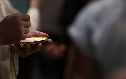 The mystery of faith. Close up of a Catholic priest serving mass and the bread of holy eucharist at communion. Darkened background but lightened host or eucharistic wafer and brass vessel