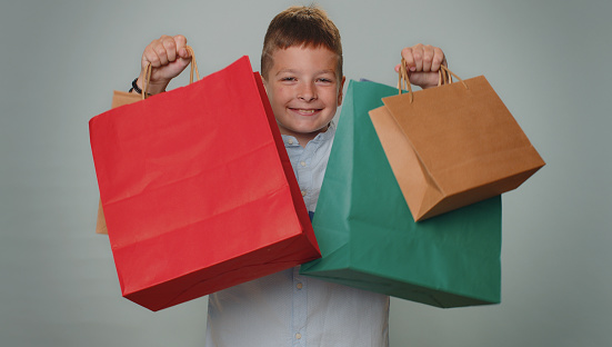 Toddler boy showing shopping bags advertising discounts, smiling looking amazed with low prices, shopping on Black Friday holidays. Young teenager children child kid isolated on gray studio background