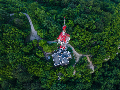 Drone view of telecommunications tower surrounded by green forest trees with cityscape before the rain on background. Technology and Global Business.