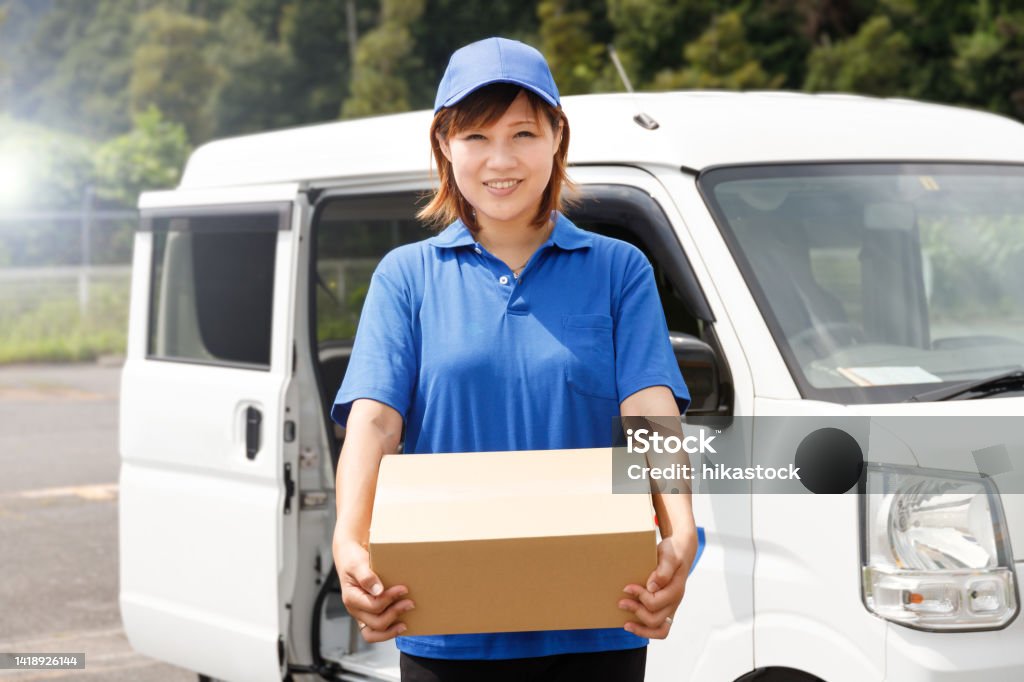 Woman in blue polo shirt and hat carrying cardboard boxes. She works as a mover and courier. Delivering Stock Photo