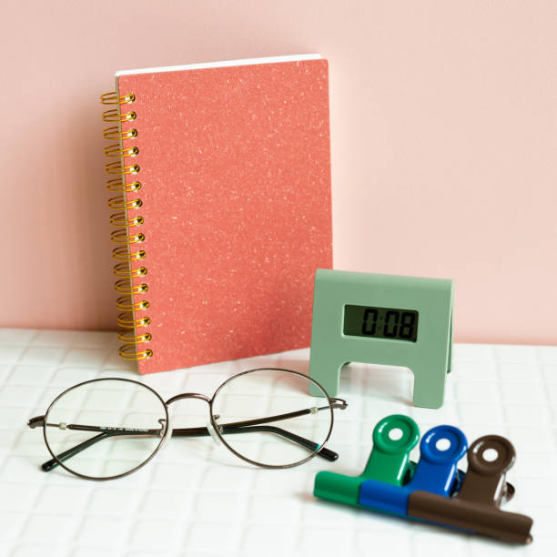 istock Office desk table with notebook, eyeglasses, clock, clip, supplies on white desk. pink wall background 1418944838