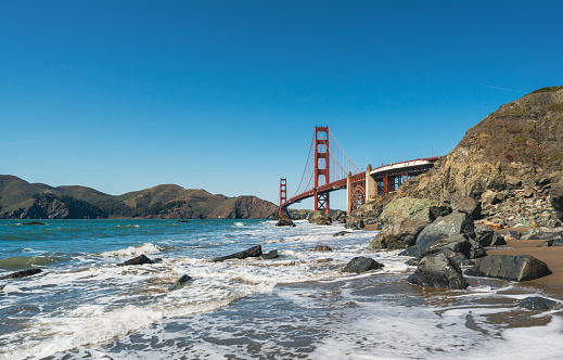 The Golden Gate Bridge in San Francisco seen a sunny morning at the beach in California, USA. Waves from the Pacific Ocean splashing towards the rocks.