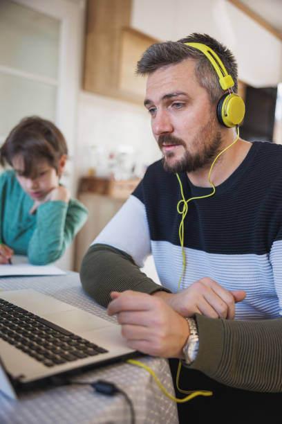 istock Man with headphones on watching content on laptop, his son doing homework 1418948766