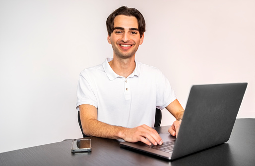 Portrait of a confident young businessman sitting at table with laptop and mobile phone looking at camera and smiling in office