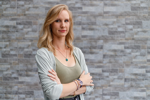 Portrait of confident caucasian female entrepreneur with arms crossed and blond hair posing against brick wall outside office
