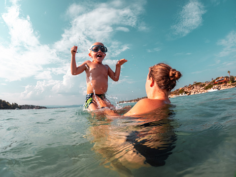 Little caucasian boy and his mother playing and jumping in the water at the beach. They are laughing and having a wonderful family time while on vacations in Greece.