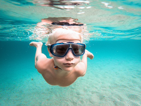 Funny photo of a cute caucasian kid swimming underwater, wearing a snorkeling mask. He is in a beautiful beach with clear turquoise color sea.
