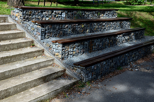 grandstand in the park by a concrete staircase made of gabion baskets with gray stones arranged inside. the seating areas of the benches are steps made of planks.