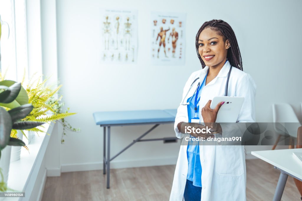 Portrait of female African American doctor standing in her office at clinic. Portrait of female African American doctor standing in her office at clinic.  Portrait Of Smiling Black Female Doctor In White Coat Posing With Folded Arms Over Light Background. 20-24 Years Stock Photo