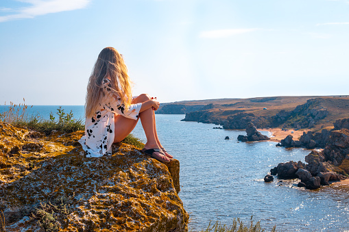 A woman traveler with long hair sits on the edge of a mountain and looks at the seascape. Travel and tourism.