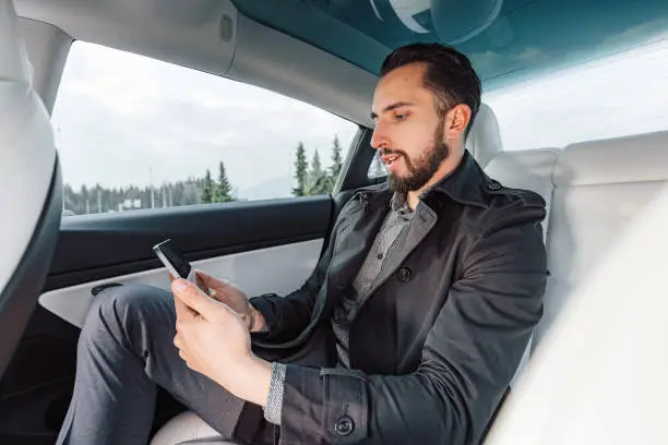 Caucasian young successful man seriously talking on the smartphone while sitting in the back seat of a driving car