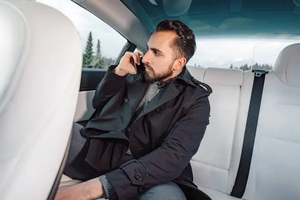 Caucasian young successful man seriously talking on the smartphone while sitting in the back seat of a driving car