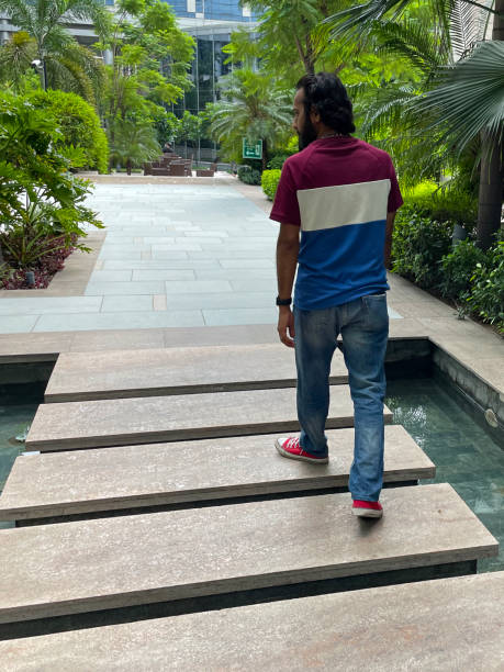 istock Image of Indian man walking over rectangular stepping stones crossing formal, tiled garden pond, paved pathways, flowerbed shrubs, water reflection, focus on foreground 1418990900