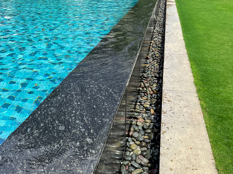 Stock photo showing close-up view of public swimming pool with blue mosaic tiles in shades of blue and low, marble effect surrounding wall and pebbles besides realistic, fake, green grass.