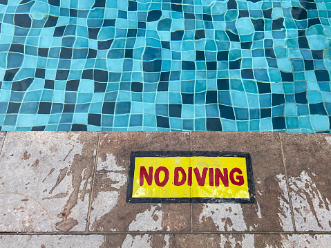 Stock photo showing close-up, elevated view of public swimming pool with blue mosaic tiles in shades of blue. A red and yellow no diving warning sign is painted on the surrounding patio area for health and safety to prevent accidents and potential drowning hazard for swimmers.