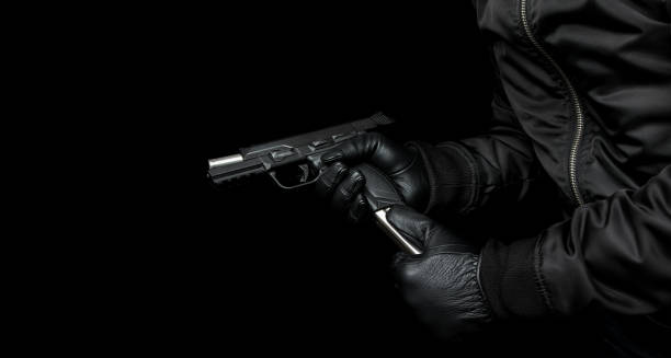 검은 재킷과 검은 장갑을 낀 남자가 권총을 손에 들고 다시 장전합니다. 손에 언로드 된 무기. 어두운 배경. - hit man 뉴스 사진 이미지