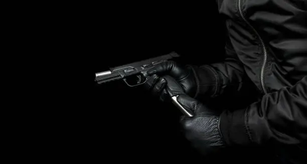 A man in a black jacket and black gloves holds a pistol in his hands and reloads it. Unloaded weapon in hand. Dark background.