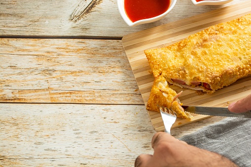 cutting brazilian snack pastel, over wooden table with sauce, pepper, ketchup also fork and knife. traditional pastry