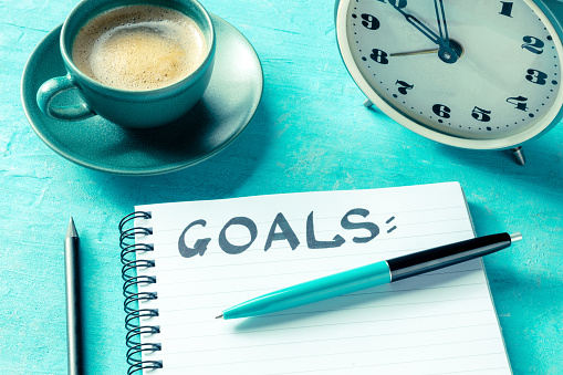 Goal setting. A notepad with the handwritten word Goals, with a pen, an alarm clock, and a cup of coffee. The concept of resolutions and lists, on a blue background