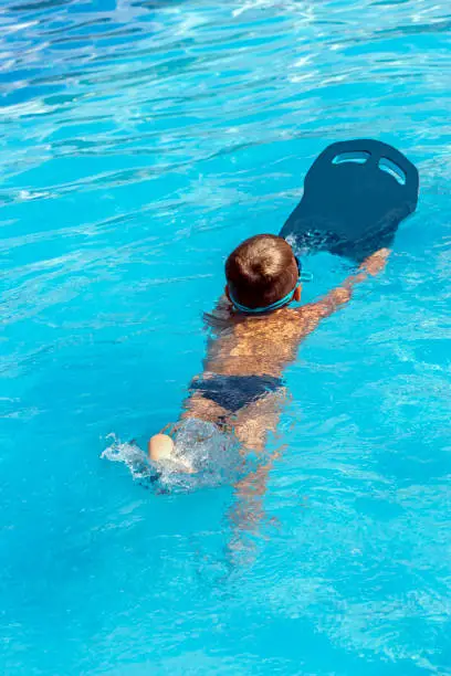 Little boy beginner swimmer learn to swim in pool. Swim training for kid in water pool by using surf and swimming kickboard . Swimming lessons and water safety education for children