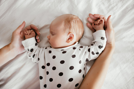 Newborn baby sleeps calmly and sweetly on the bed holding his mother's hands