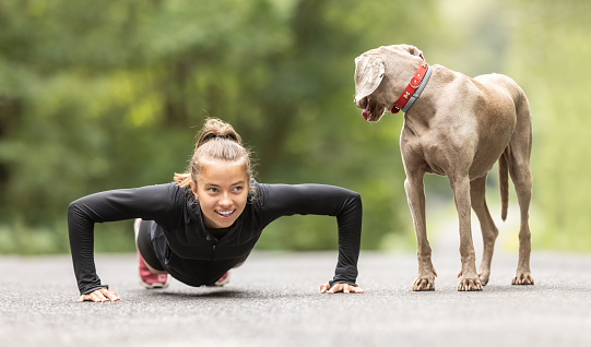 Young woman doing pushups outdoors smiles at her dog observing her.
