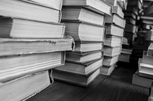 Stacks of books on a wooden table. Black and white photograph of books collected in stacks on the table. Several stacks of books stand side by side on the floor. Lots of books in one place.