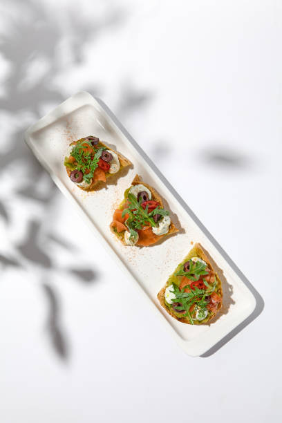 istock Aesthetic composition with salmon bruschetta on white background with shadows from flowers. Italian bruschetta with salmon, avocado, cheese and olives on fine dining in summer. Elegant menu concept. 1419017756