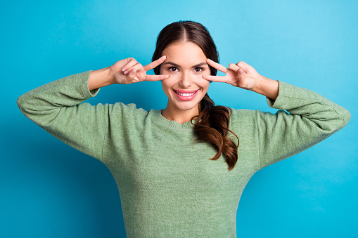 Photo of cheerful pretty girl hands fingers show v-sign near eyes toothy smile isolated on blue color background.