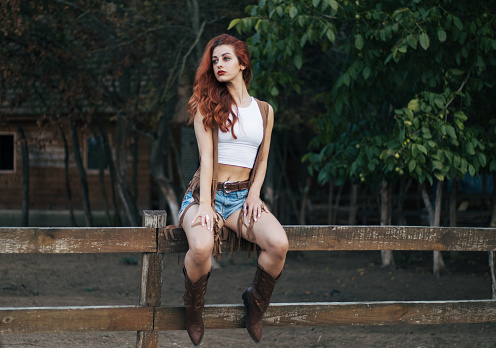 Young woman with red hair on farm posing in cowgirl outfit with energic summer vibes, With beautiful boots and horses. White crop top with shorts and brown vest.