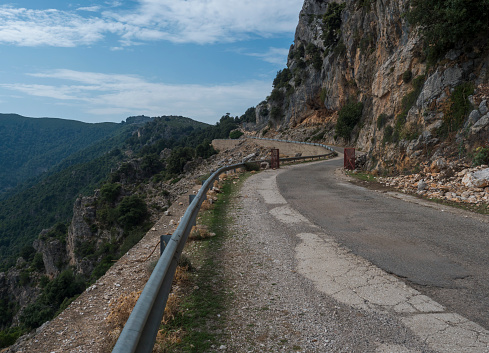 Narrow dangerous asphalt road at Genna Croce pass. at Supramonte Mountains with white limestone rocks, green hills, trees and mediterranean forest vegetation. Sardinia, Italy. Summer cloudy sky