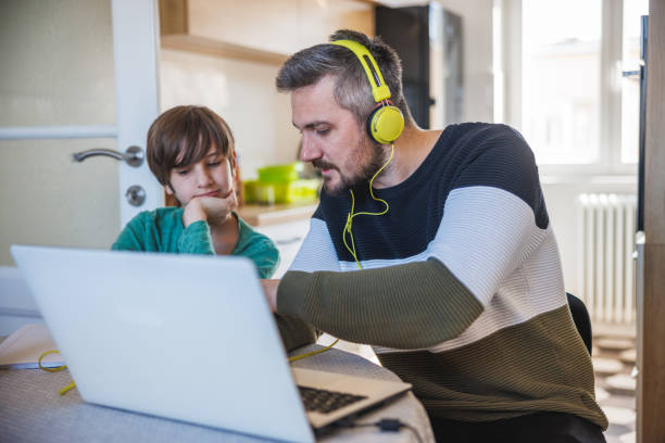 man working on laptop at home, his son sitting next to him and looking at content - inconvenience meeting business distracted imagens e fotografias de stock