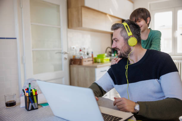 man smiling and working on laptop at home, his son distracting him standing behind and laughing - inconvenience meeting business distracted imagens e fotografias de stock