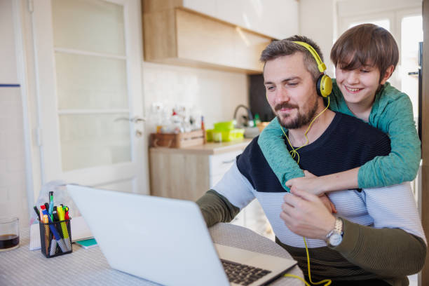 man working on laptop at home, his son embracing him from behind and smiling - inconvenience meeting business distracted imagens e fotografias de stock