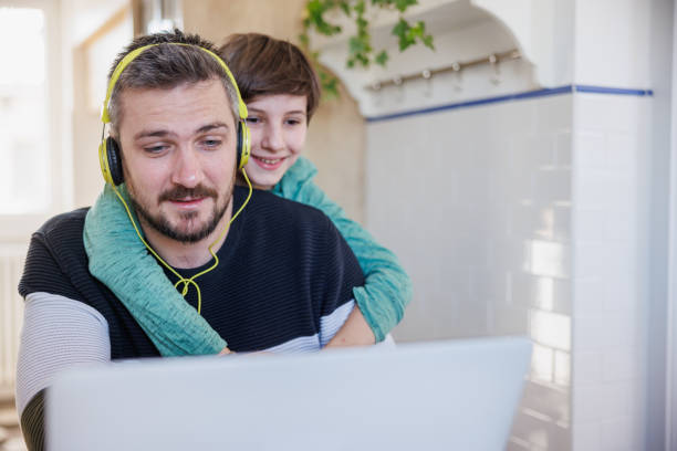 patient man working on laptop at home, his son embracing him from behind and smiling - inconvenience meeting business distracted imagens e fotografias de stock