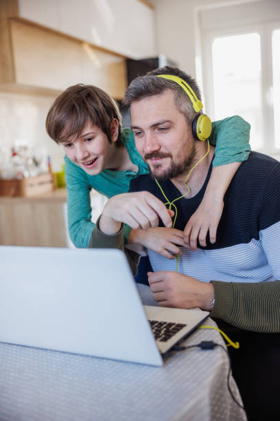 man smiling and working on laptop at home, his son embracing him from behind and laughing - inconvenience meeting business distracted imagens e fotografias de stock