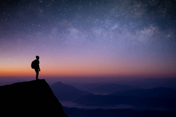 Silhouette of young traveler with backpack standing and watched the star, milky way and night sky alone on top of the mountain. He enjoyed traveling and was successful when he reached the summit. stock photo