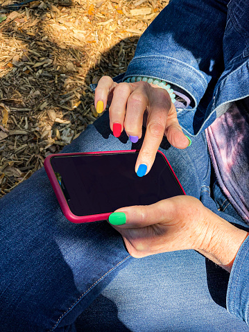 A 58-year-old caucasian female is touching the screen of her cell phone. Her fingernails are colored in primary colors. There is a map present on the cell phone. Copy space is available in the blackened cell phone screen
