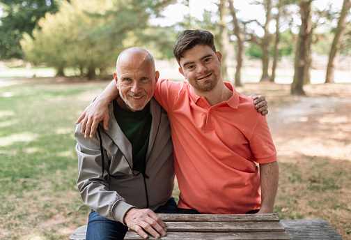 A happy senior father with his young son with Down syndrome embracing and sitting in park.