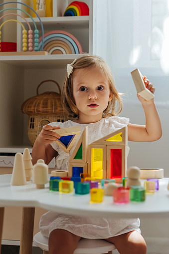 A little girl playing with wooden blocks on the table in playroom.  Educational game for baby and toddler in modern nursery. The kid builds a tower from wooden rainbow stacking blocks.