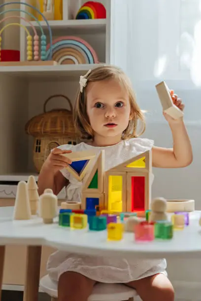 Photo of A little girl playing with wooden blocks on the table in playroom.  Educational game for baby and toddler in modern nursery. The kid builds a tower from wooden rainbow stacking blocks.