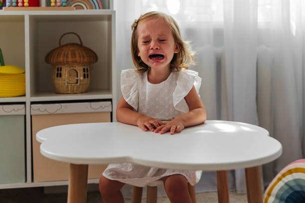A little girl cries and is naughty sitting at the table in the nursery. Children's tantrums, tears and discontent. Problems of kid's upbringing.