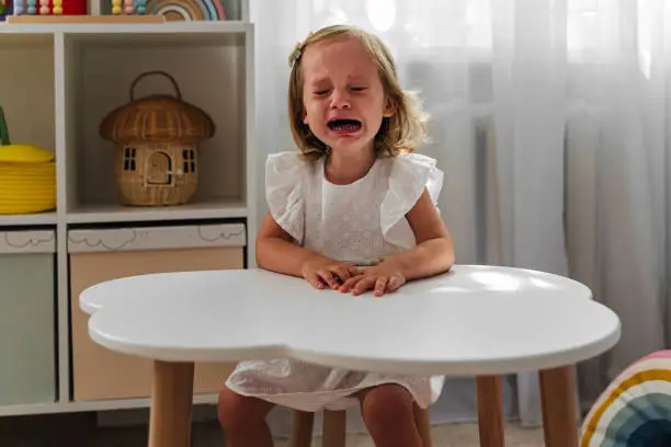 Photo of A little girl cries and is naughty sitting at the table in the nursery. Children's tantrums, tears and discontent. Problems of kid's upbringing.