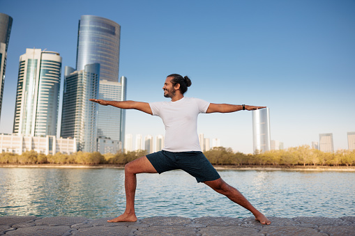 Man doing yoga on mat by the sea in Abu Dhabi, standing in lunge with arms outstretched in Warrior 2 Virabhadrasana II pose, city skyline behind