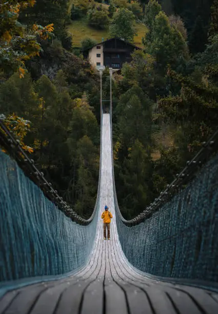 Photo of Young traveler man dressed in yellow jacket crosses hiking on an impressive wooden and metal bridge in the village of Goms in the Swiss alps